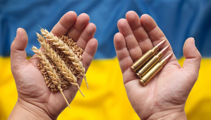 Image d'un homme tenant du blé et des balles dans ses mains en étant au dessus d'un drapeau ukrainien.